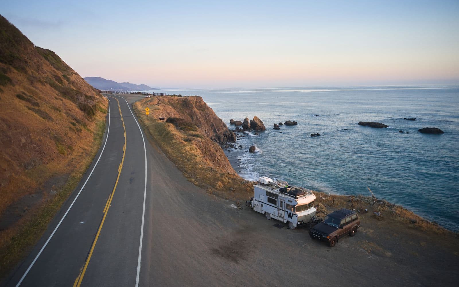 A camper van parked above a coastal highway at sunset in California